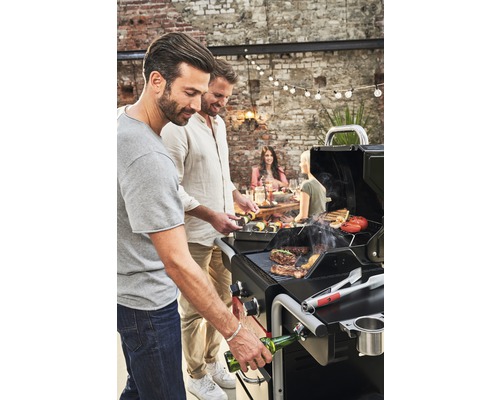 Zwei Männer grillen Fleisch und Gemüse auf einem Gasgrill im Freien, während andere am Tisch sitzen.