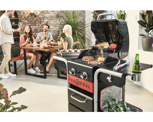 Grillszene mit Gas-Kohle-Grill, Tisch und Personen im Freien.