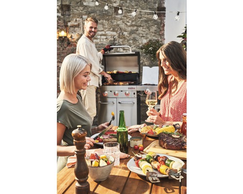 Szene mit einem Grillabend im Freien, bei dem gegrilltes Essen auf einem Tisch serviert wird und Personen zusammensitzen