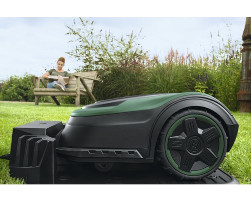 Bosch Mähroboter auf Rasen mit Frau auf Gartenbank im Hintergrund