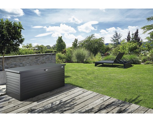 Gartenszene mit einer Gerätebox aus Metall auf einer Holzterrasse und einer Sonnenliege auf dem Rasen