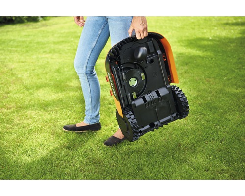 Eine Person trägt einen Rasenmähroboter über eine Rasenfläche.