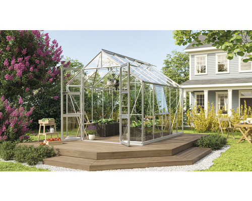 Gewächshaus Vitavia Olymp 8300 Einscheibensicherheitsglas 3 mm 257 x 324 cm silber Gewächshaus mit Glasfront auf einer Holzterrasse in einem Garten mit Pflanzen und einem Haus im Hintergrund.