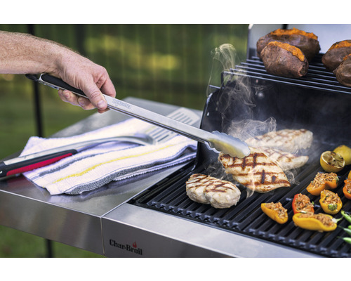 Eine Grillzange wendet Hähnchenfleisch auf einem Grill, zusammen mit Kartoffeln und gefüllten Paprika.