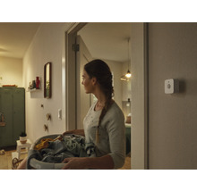 Eine Frau trägt einen Wäschekorb durch einen Flur, an der Wand ist ein Smart Home Bewegungsmelder montiert.