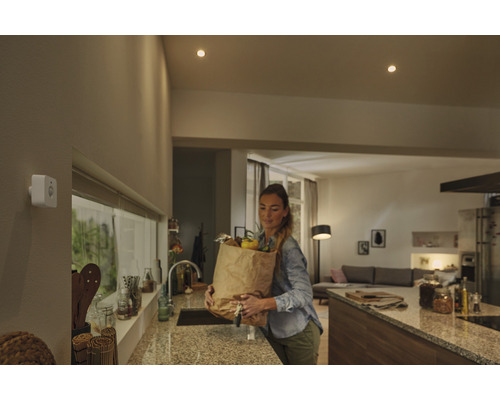 Weißer Bewegungsmelder an einer Wand in einer modernen Küche mit einer Frau, die eine Einkaufstüte trägt.