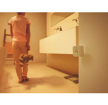 Ein weißer quadratischer Bewegungsmelder an der Wand in einem schwach beleuchteten Badezimmer mit einer Person, die einen Teddybären hält.