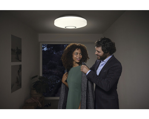 Szene mit einer LED Deckenleuchte in einem Flur, unter der sich ein Paar befindet, wobei der Mann der Frau den Mantel anzieht.