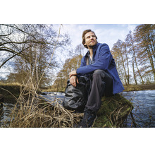Ein Mann sitzt mit Rucksack an einem Bachufer in der Natur.