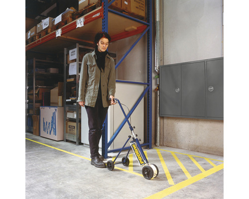 Frau zieht mit einem Markierungswagen gelbes Markierungsband auf den Boden eines Lagers.