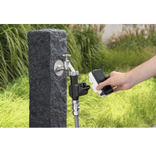 Gartenszene mit Wasserhahn an einem Steinpostament und einer Person, die ein Bewässerungsgerät anschließt