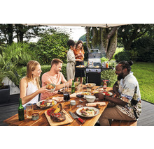 Eine Gruppe von Personen grillt und isst an einem Holztisch im Garten mit einem Gasgrill im Hintergrund.