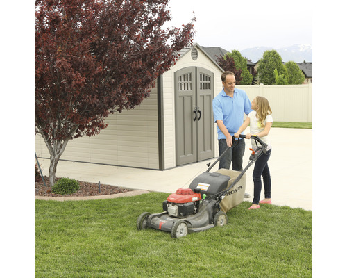 Vater und Tochter mähen Rasen mit einem Rasenmäher vor einem Gartenhaus.