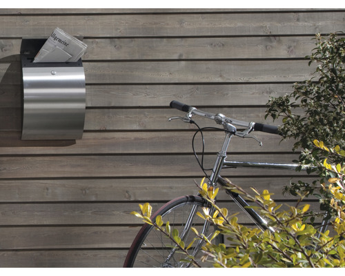 Zeitungsfach aus Edelstahl mit Zeitung an einer Holzwand, daneben ein Fahrrad