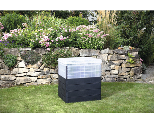 Komposter aus Kunststoff im Garten vor einer Steinmauer und Blumenbeeten