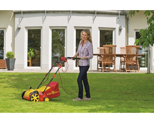 Frau benutzt einen gelb-roten Rasenmäher der Marke Wolf Garten auf einer Rasenfläche vor einem Haus.