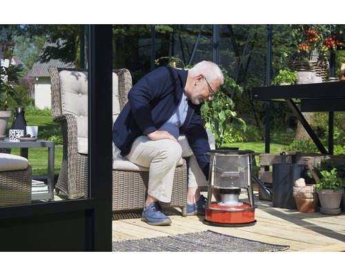 Mann bedient eine Petroleumheizung in einem hellen Gewächshaus mit Möbeln und Pflanzen.