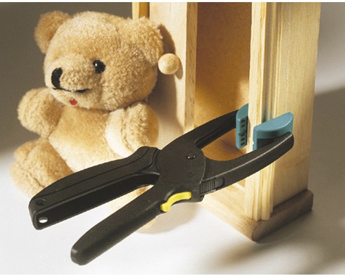 Federzwinge beim Fixieren eines Holzstücks an einem Miniaturschrank, mit einem Teddybär im Hintergrund
