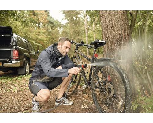Mann reinigt Fahrrad mit Wasserschlauch im Wald. Im Hintergrund ist ein Auto zu sehen.
