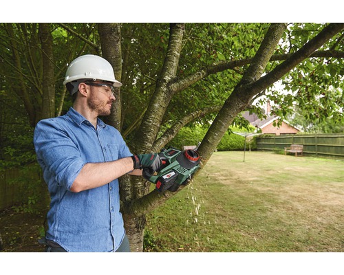 Mann mit Schutzhelm und Schutzbrille schneidet einen Baumast mit einer Akku-Astsäge im Garten.