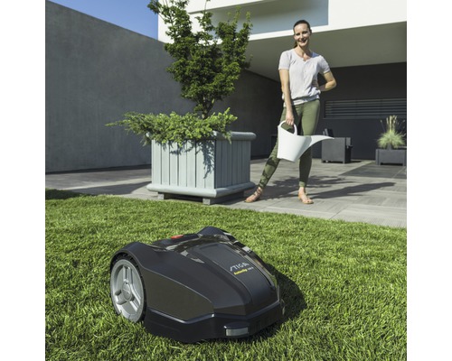 Schwarzer Rasenmähroboter auf Rasen mit Frau im Hintergrund, die eine Gießkanne hält