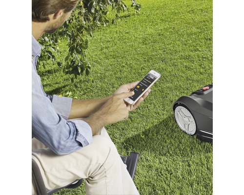 Eine Person bedient einen Rasenmähroboter über eine Smartphone-App im Garten.