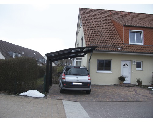 Ein Carport aus Metall vor einem Wohnhaus mit gepflasterter Auffahrt und einem geparkten Auto.