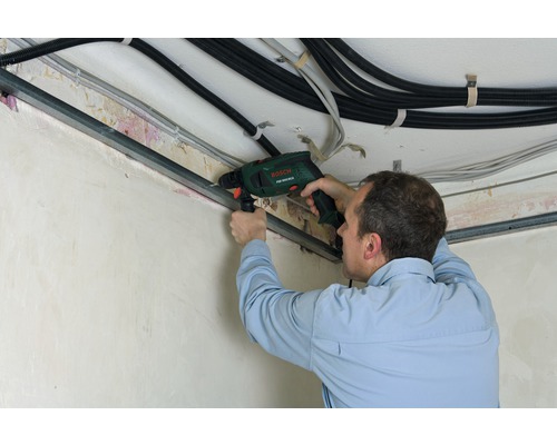 Mann bohrt mit einer Schlagbohrmaschine in eine Metallschiene an der Decke. Kabel sind ebenfalls sichtbar.