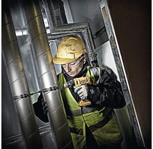 Ein Handwerker mit Schutzhelm und Schutzweste bohrt mit einem Akkubohrer in eine Rohrleitung.