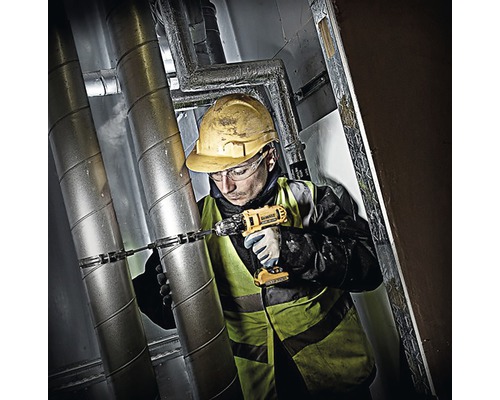Ein Handwerker mit Schutzhelm und Schutzweste bohrt mit einem Akkubohrer in eine Rohrleitung.