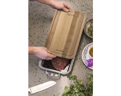 Person legt Holzschneidebrett auf eine Edelstahlform mit Fleisch, daneben liegen Zutaten und ein Messer.