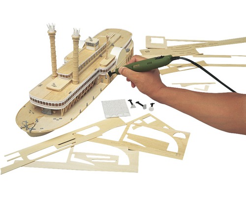 Modellbauszene mit einem Schaufelraddampfer aus Holz, verschiedenen Holzteilen und einem Multifunktionswerkzeug in einer Hand.