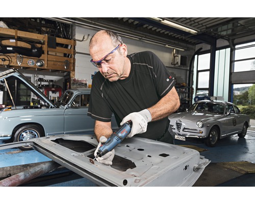 Mann arbeitet mit einem Multifunktionswerkzeug an einer Autotür in einer Werkstatt mit Oldtimern.
