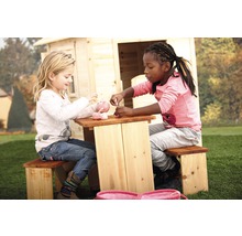 Zwei Kinder sitzen an einem Picknicktisch aus Holz und spielen mit einem Teeservice vor einem Spielhaus.