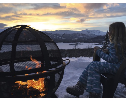 Szene mit Feuerstelle im Freien, Kind sitzt im Stuhl, Schnee und Berglandschaft im Hintergrund