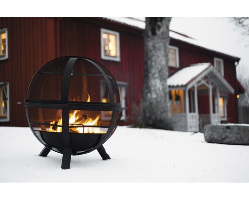 Kugelgrill aus Metall mit Feuer vor rotem Haus im Schnee