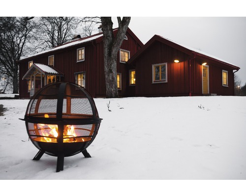 Feuerkorb aus Metall mit brennendem Feuer im verschneiten Garten vor einem Haus.