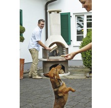 Mann legt Holz in einen Gartengrill während ein Hund zusieht.