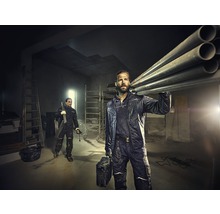 Zwei Handwerker in Arbeitskleidung transportieren Rohre und Werkzeug in einem hellen Raum mit Leitern.