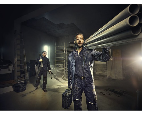 Zwei Handwerker in Arbeitskleidung transportieren Rohre und Werkzeug in einem hellen Raum mit Leitern.