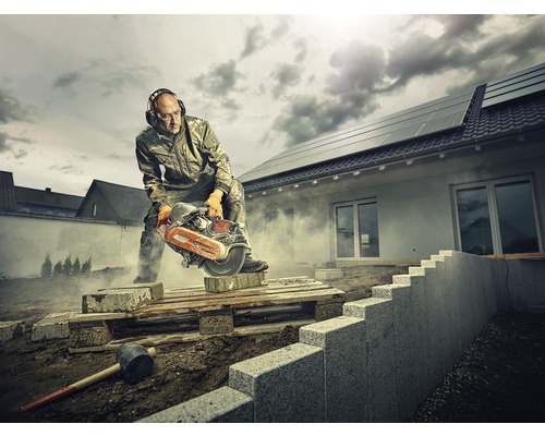 Ein Mann mit Gehörschutz und Schutzbrille schneidet einen Stein mit einer Steinsäge auf einer Baustelle.