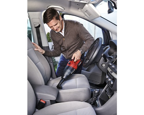 Mann saugt mit einem Akku-Handsauger den Innenraum eines Autos. Der Staubsauger ist rot und schwarz.