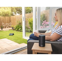 Frau mit Tasse und Amazon Echo neben Rasenmähroboter im Garten