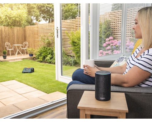 Frau mit Tasse und Amazon Echo neben Rasenmähroboter im Garten