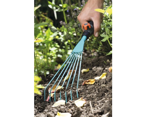 Hand mit Gartenrechen beim Entfernen von Laub aus dem Gartenbeet