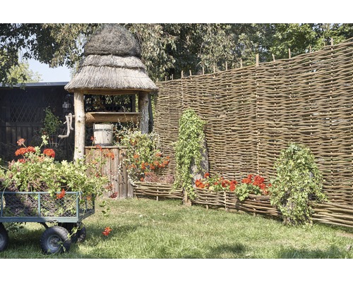Szene mit Gartenbrunnen, Weidenzaun, Blumenwagen und bepflanzten Weidenkästen
