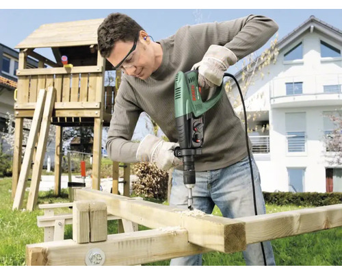 Mann bohrt mit einer Schlagbohrmaschine Löcher in ein Holzbrett im Garten.