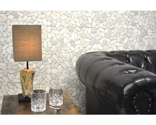 Szene mit einer Tischlampe auf einem Holztisch, zwei Gläsern, einem Flaschenöffner und einem Ledersofa vor einer Wand mit Kieselsteinen.