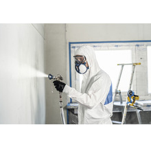Mann mit Schutzanzug, Schutzbrille und Maske beim Sprühen einer Wand mit einer Farbspritzpistole