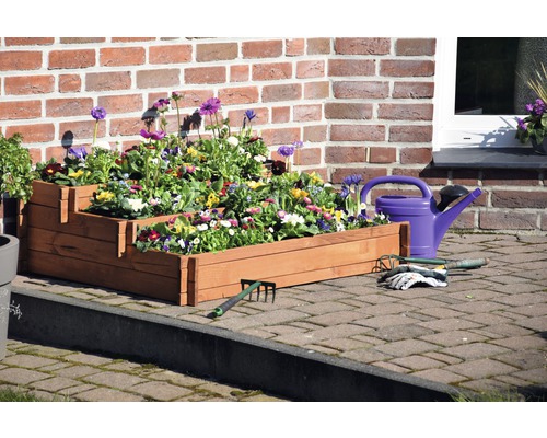 Gestufter Pflanzkasten aus Holz mit Blumen, Gießkanne und Gartengeräten vor einer Backsteinmauer